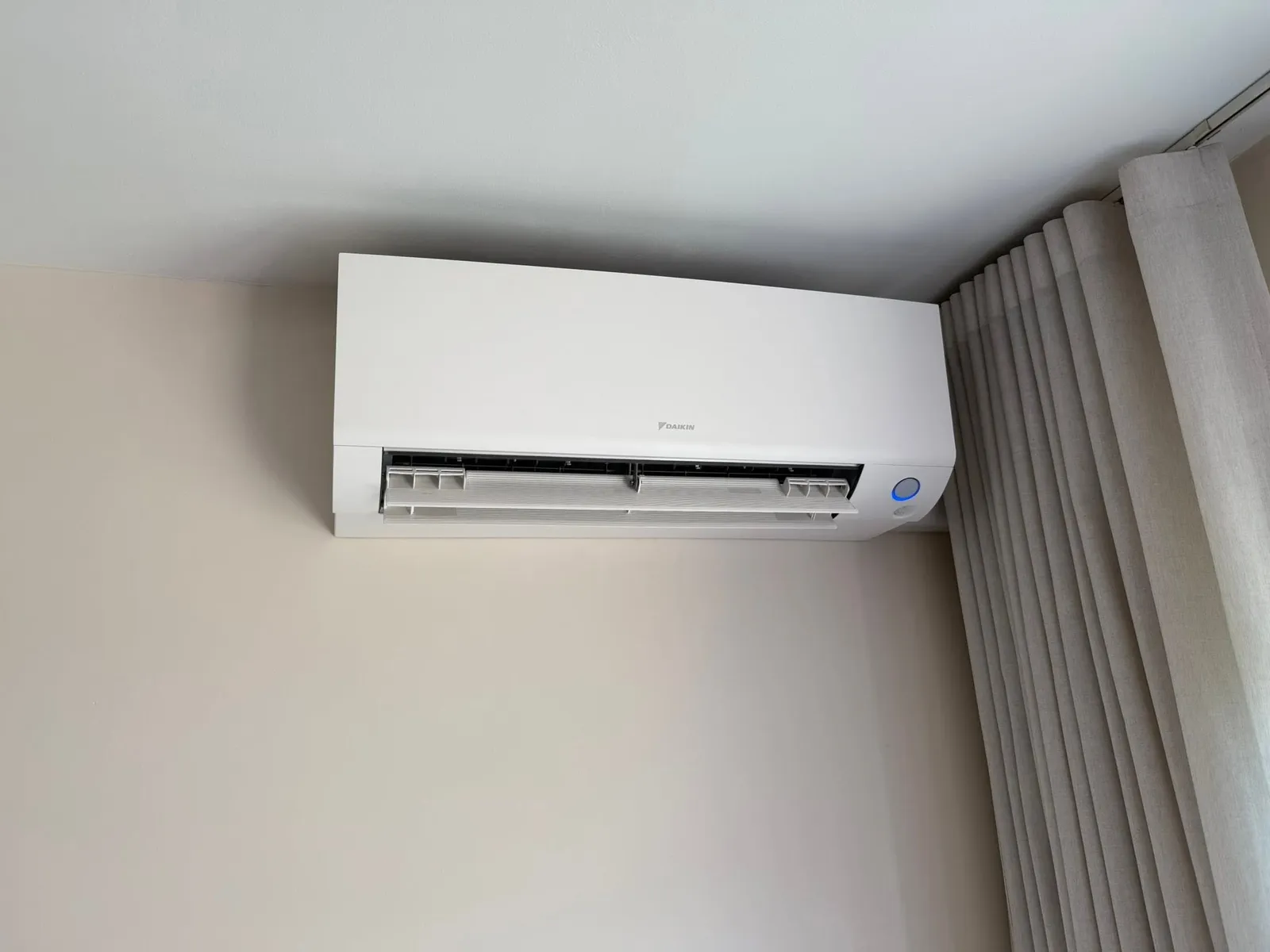 Wit Daikin airco-binnendeel hoog tegen de wand gemonteerd, naast de gordijnrail