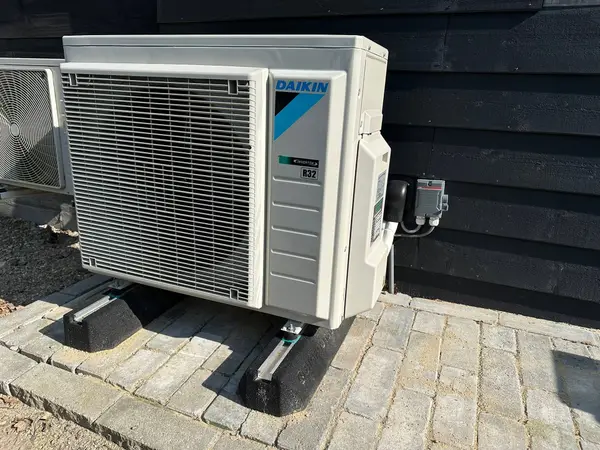 Buitenunit op klinkers tegen een donkere houten gevel